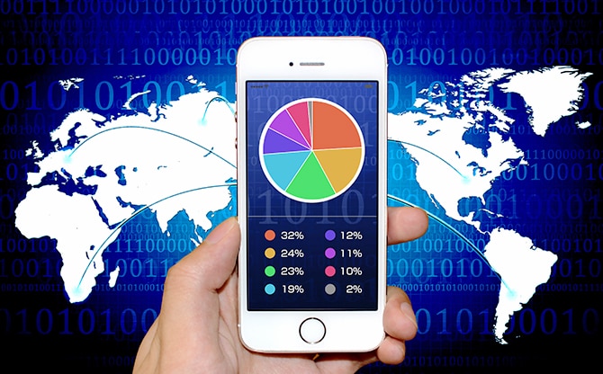 手に持つ円グラフが表示された白いスマートフォンが世界地図の各国と線でつながっているイメージ